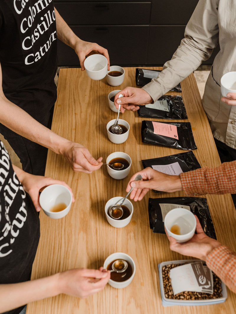 People around a wooden table tasting coffee from small white cups.